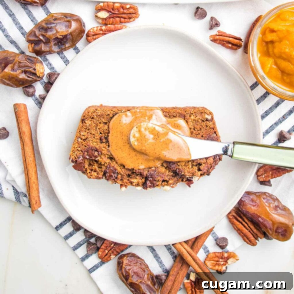 Slice of pumpkin bread on a white plate and a dollop of almond butter with a jewel green spreader. Dates, cinnamon sticks, pecans and chocolate chips are scattered around the plate.