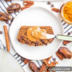 Slice of pumpkin bread with a dollop of almond butter on a white plate. There are dates, cinnamon sticks, pecans and chocolate chips strewn around the sides of the plate.