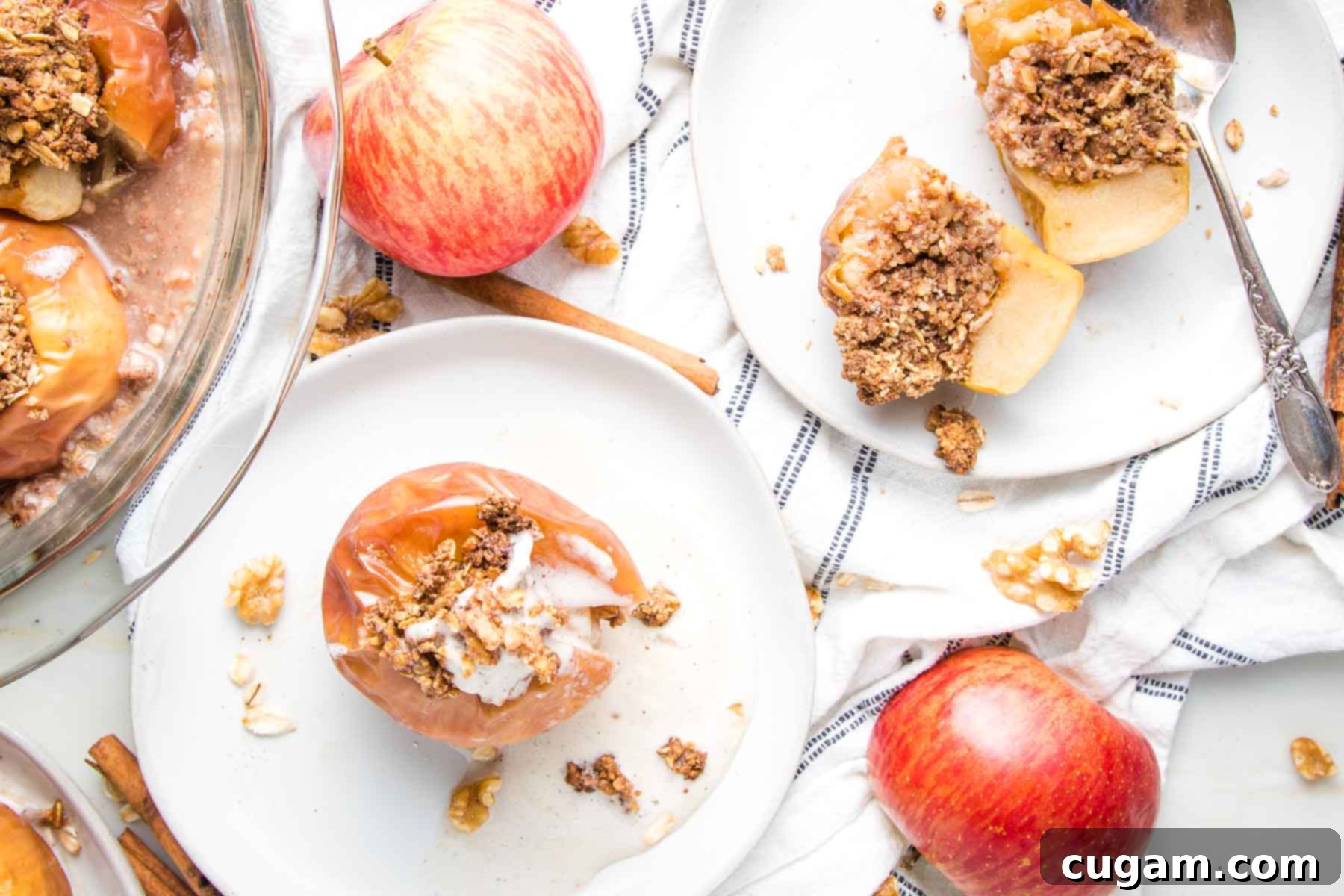 Edge of a glass pie dish filled with baked apples. A whole baked apple on a white rimmed plate with a small scoop of vanilla ice cream inside and another white plate with a whole baked apple cut in half so you can see the oatmeal cinnamon filling inside. There are whole red apples and crumbs of oats and walnuts strewn around along with a navy striped tea towe.