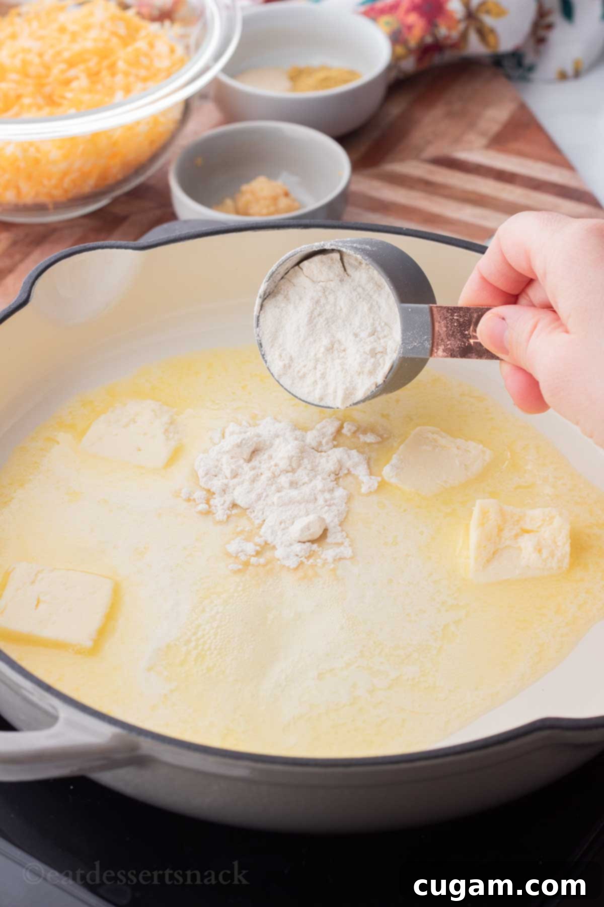 A hand holding a gray measuring cup, precisely pouring all-purpose flour into a sizzling frying pan filled with bubbling melted butter.