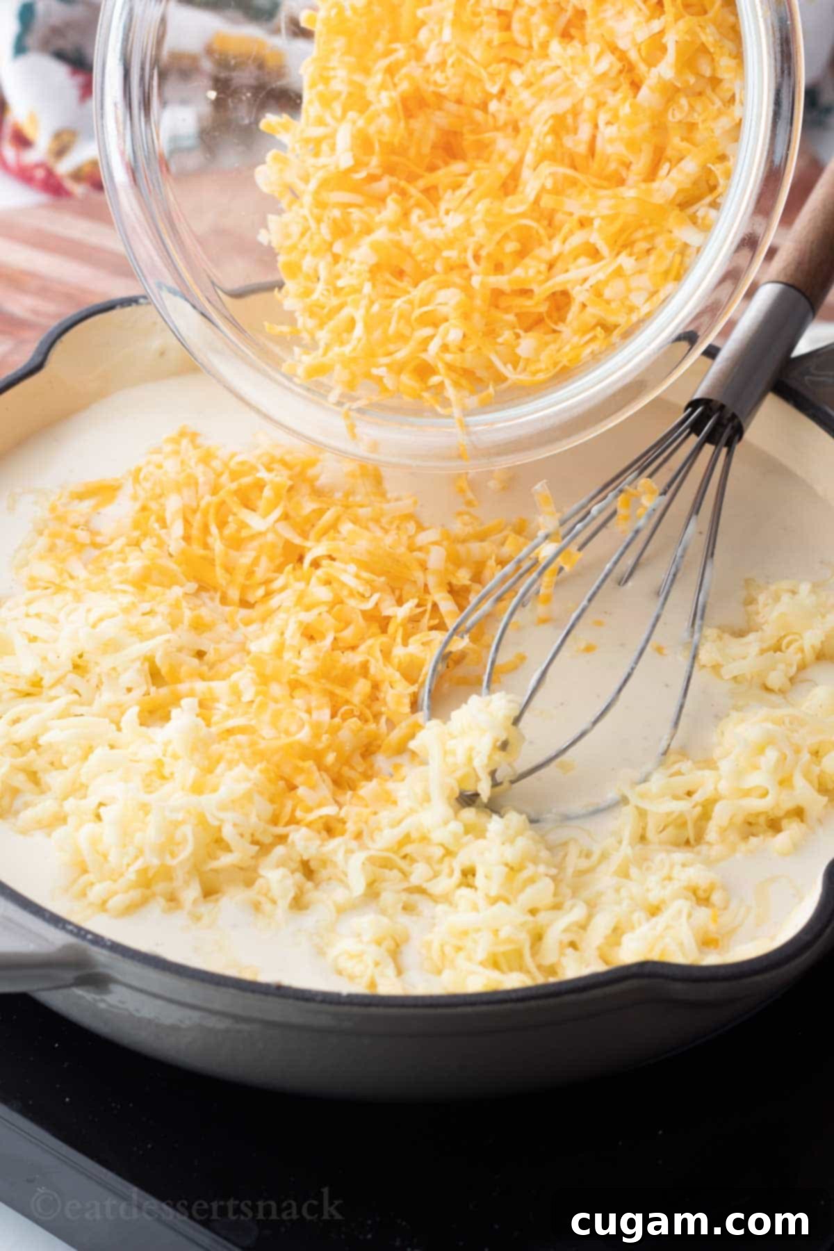 A glass bowl brimming with freshly grated cheese being poured into a large frying pan containing the creamy milk sauce, with a wire whisk poised.