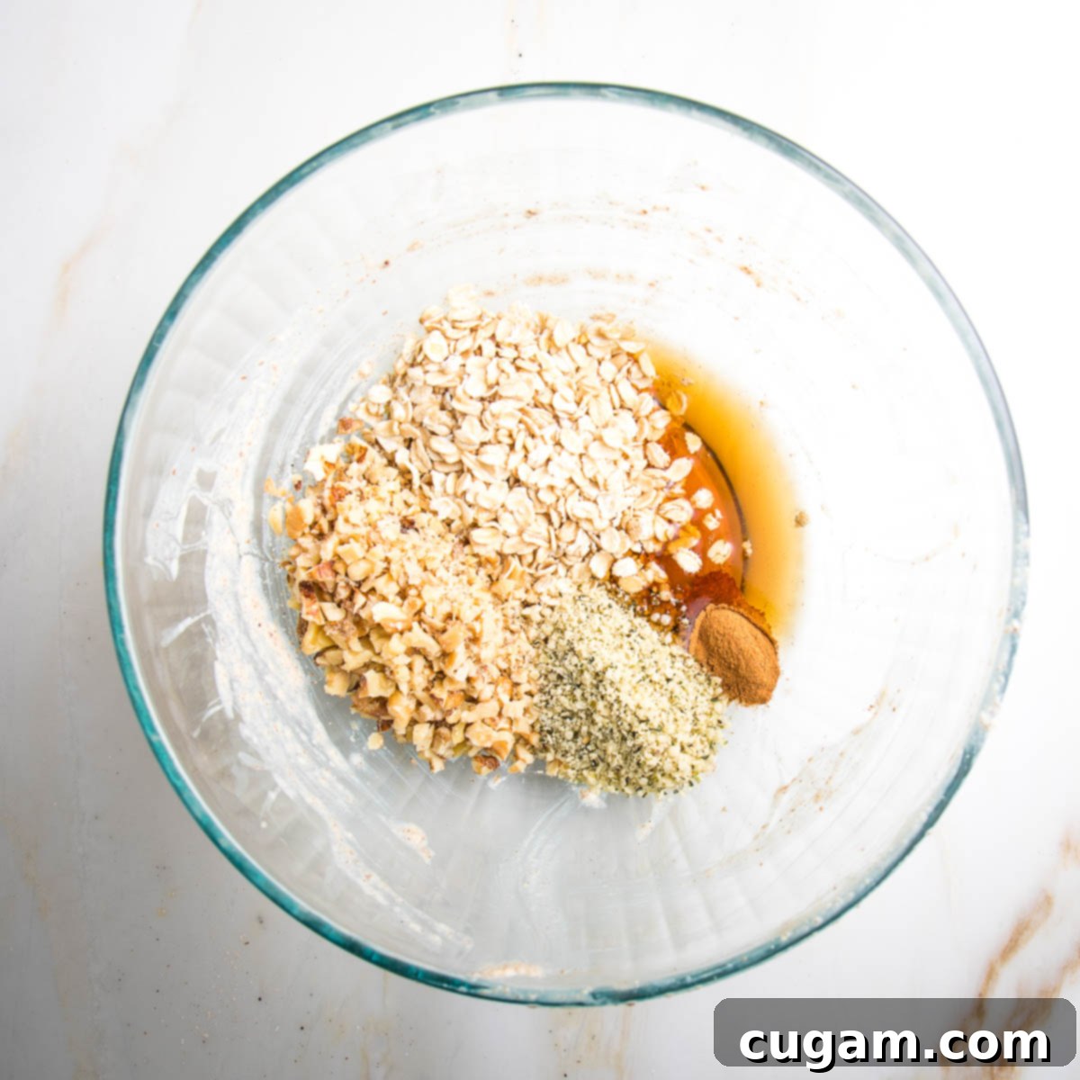 The previously used large glass mixing bowl now contains all the topping ingredients: old-fashioned oats, chopped walnuts, hemp seeds, cinnamon, and maple syrup, ready to be combined.