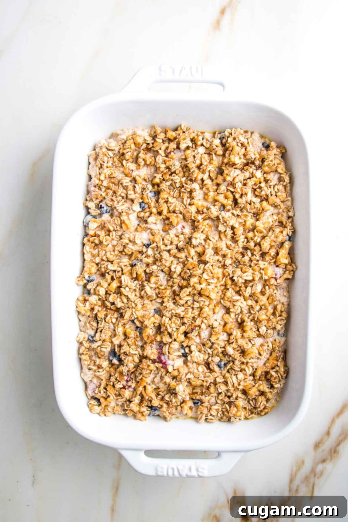 A white baking dish with the berry and oatmeal mixture now fully covered by the thick, crunchy topping, evenly distributed across the surface.