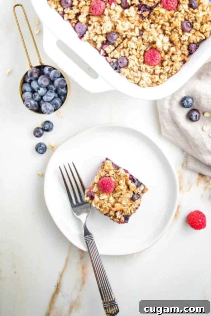 A tempting square of vegan berry baked oatmeal resting on a white plate, adorned with fresh raspberries and blueberries. The main baking dish, filled with more baked oatmeal squares, sits beside it, creating an inviting breakfast scene.