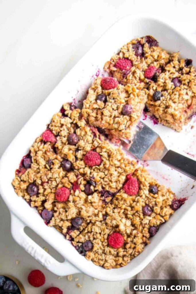 A white rectangular baking dish filled with baked oatmeal, topped with a crunchy layer of nuts and fresh berries. A spatula is gently scooping out a perfect square portion, highlighting the moist interior and appealing texture.