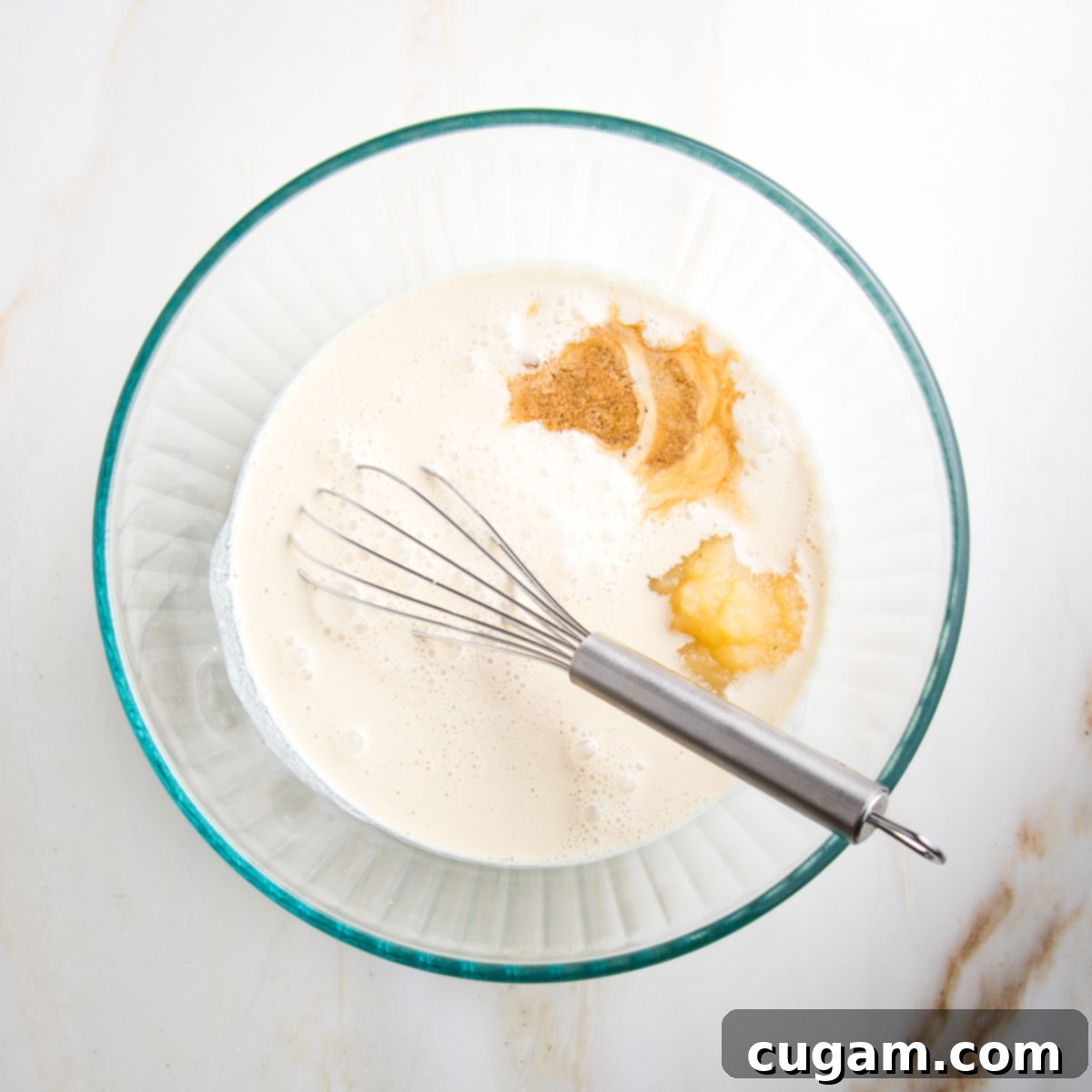 A clear glass mixing bowl containing a creamy liquid mixture of plant milk, applesauce, and vanilla extract.