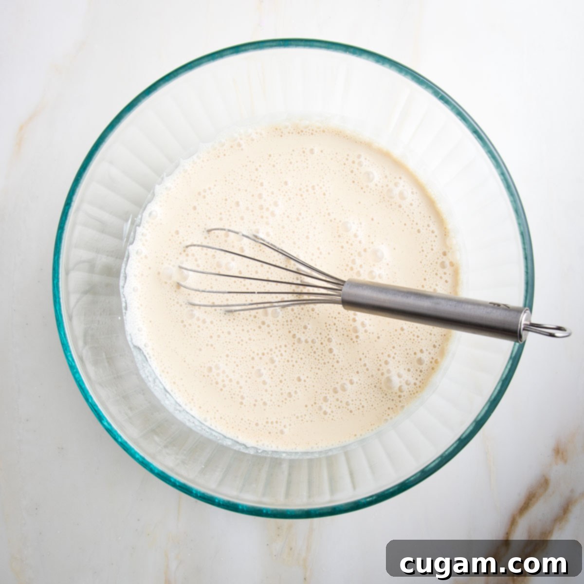 A whisk vigorously mixing a pale, creamy liquid in a glass bowl, ensuring the applesauce is fully blended into the almond milk and vanilla.