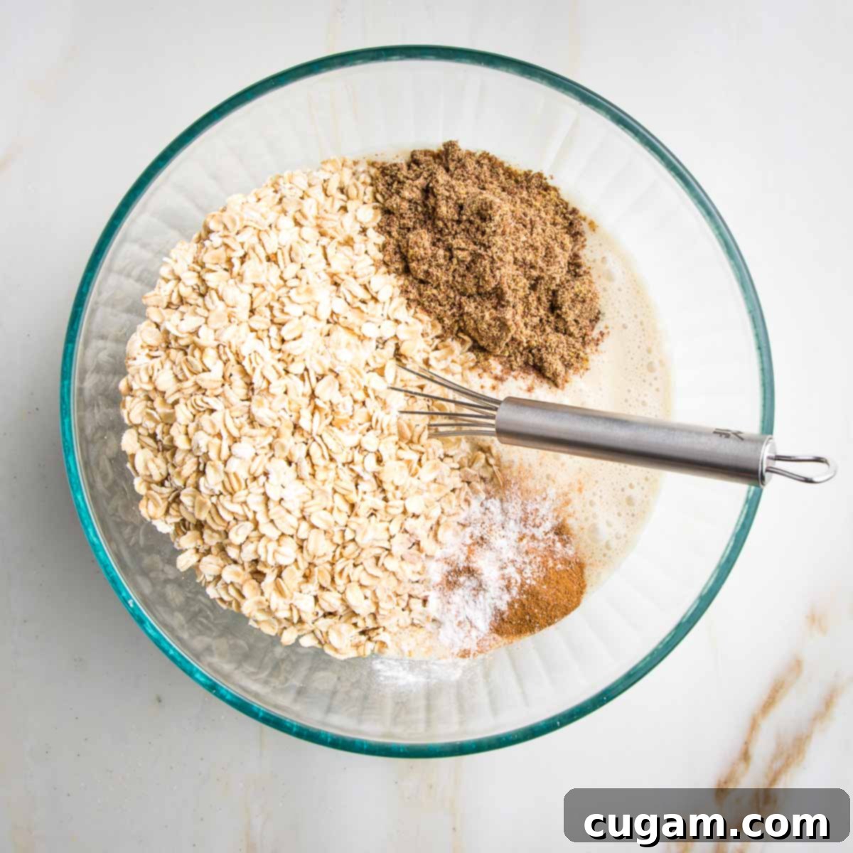 Dry ingredients, including old-fashioned oats, ground flaxseed, cinnamon, and baking soda, carefully layered on top of the creamy white liquid mixture in a bowl.