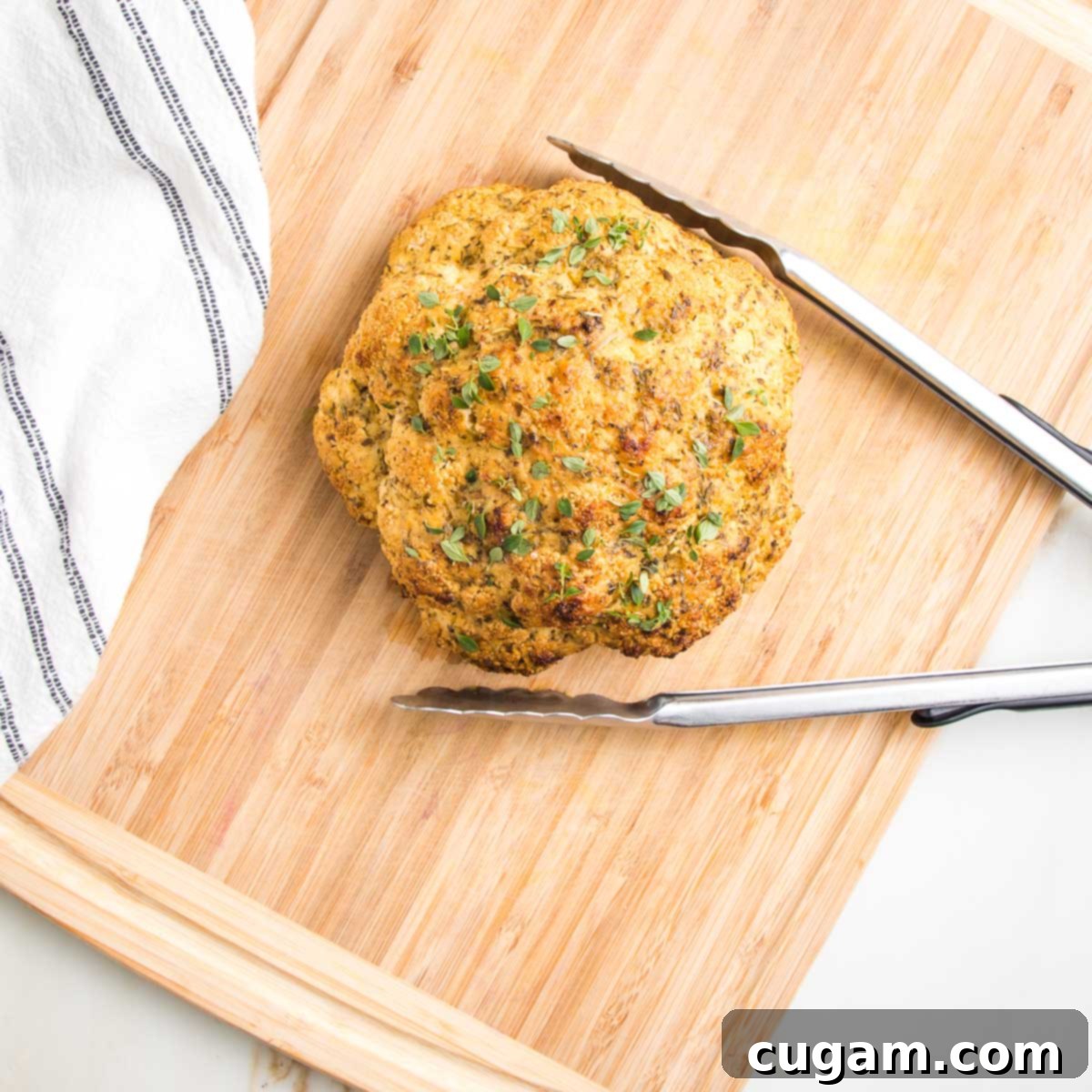 Whole roasted cauliflower with herbs on a cutting board with large tongs.