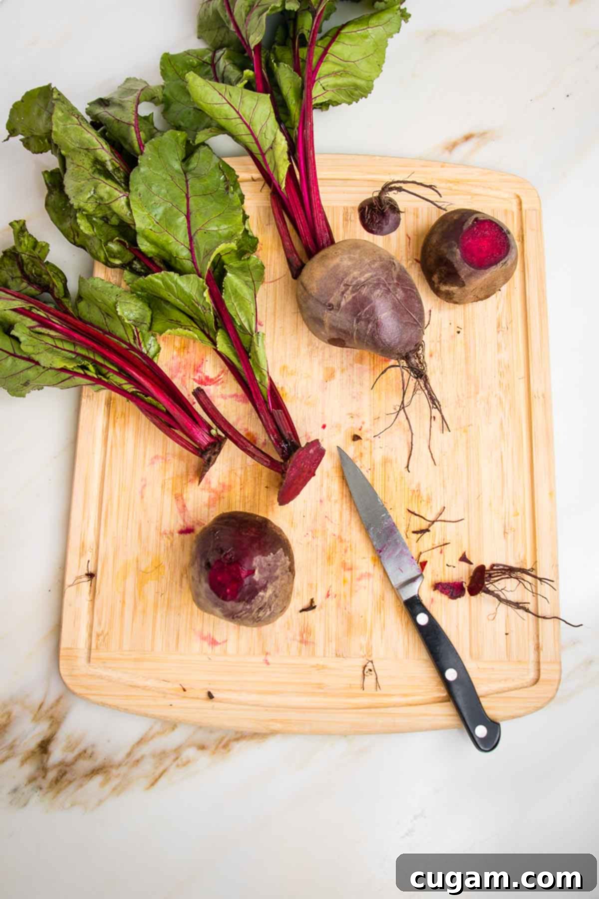 Trimming beet roots on a wooden board with a paring knife. You can see the beet greens and stems, plus three beets in various stages of trimming, ready for roasting.