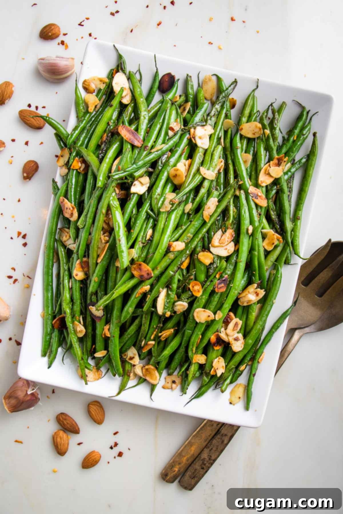 Effortless Green Beans with Toasted Almonds 2 White rectangular plate loaded with bright green beans, toasted almonds. There are cloves of garlic, sliced almonds and crushed red pepper strewn around the plate and a bronze colored large spoon and fork on the right side.