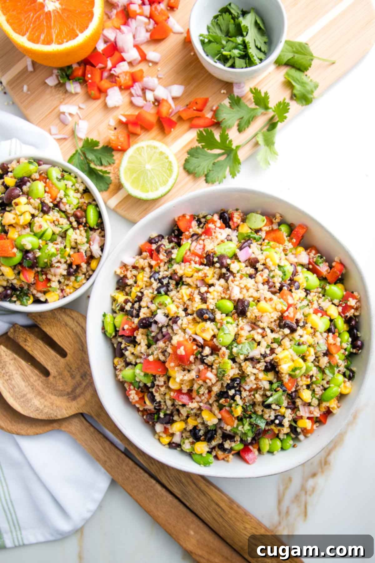 Two white bowls, one large and one small, are filled with a colorful Southwest Quinoa Salad. The salad features a mix of red peppers, green edamame, black beans, bright yellow corn, and fresh cilantro, all mixed with fluffy quinoa. In the blurred background, a wooden cutting board holds remnants of chopped red pepper, minced shallot, and fresh cilantro, alongside half an orange and half a lime. Wooden salad servers rest beside the bowls, inviting you to dig in.