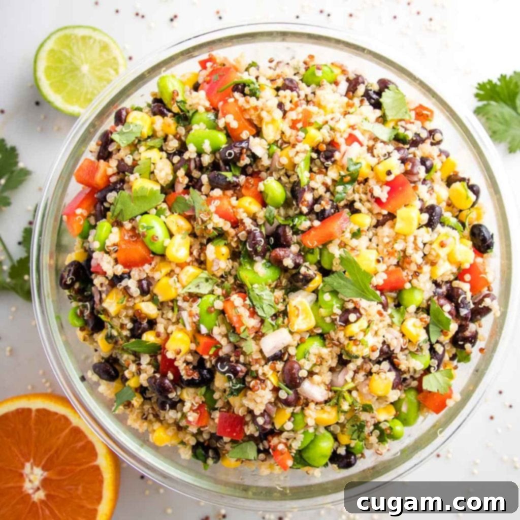 A large, clear glass bowl brimming with a vibrant, colorful quinoa salad. The salad is a lively mix of red bell peppers, green edamame, black beans, bright yellow corn, and chopped fresh cilantro, all beautifully blended with fluffy quinoa. Surrounding the bowl on a pristine white counter are half a fresh lime, half a juicy orange, and sprigs of fragrant cilantro, highlighting the fresh ingredients used in the dish.