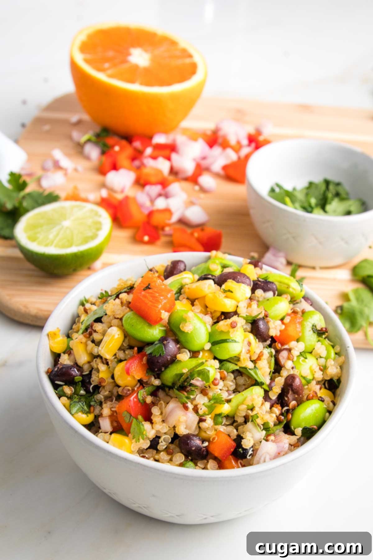 A close-up shot of a large, colorful bowl of Southwest Quinoa Salad, featuring a mix of fluffy quinoa, black beans, edamame, corn, and red bell peppers. In the softly blurred background, a cutting board displays half an orange, half a lime, chopped red peppers, minced shallots, and fresh cilantro, highlighting the fresh, vibrant ingredients used in the salad.