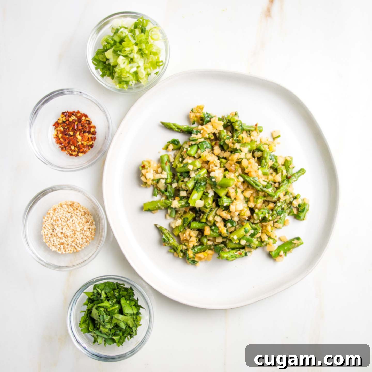 Cauliflower rice with asparagus on a rimmed white plate, surrounded on the left with small glass dishes with sliced scallions, crushed red pepper, sesame seeds and chopped fresh parsley