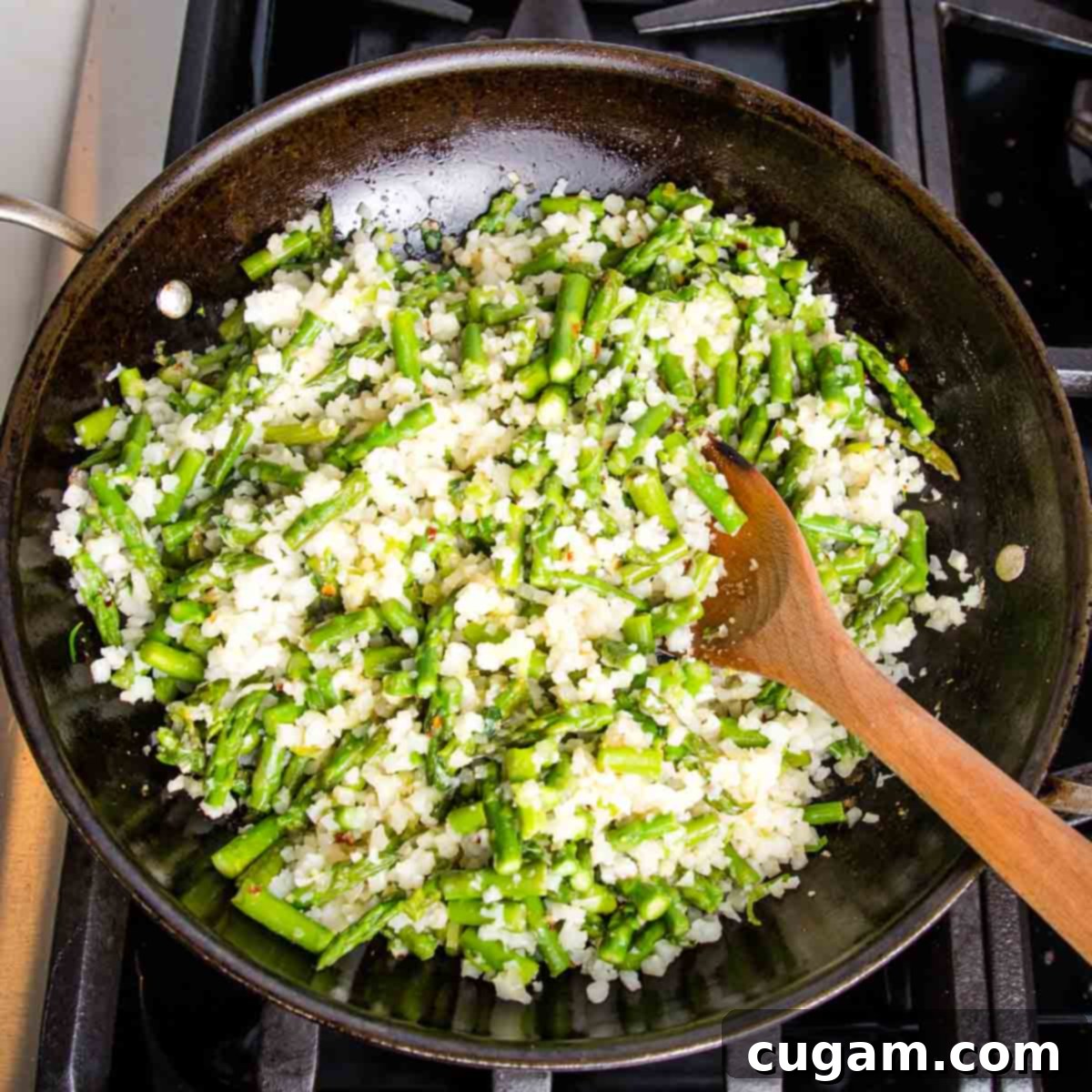 Cauliflower rice and asparagus in a skillet, stirred with a wooden spoon.