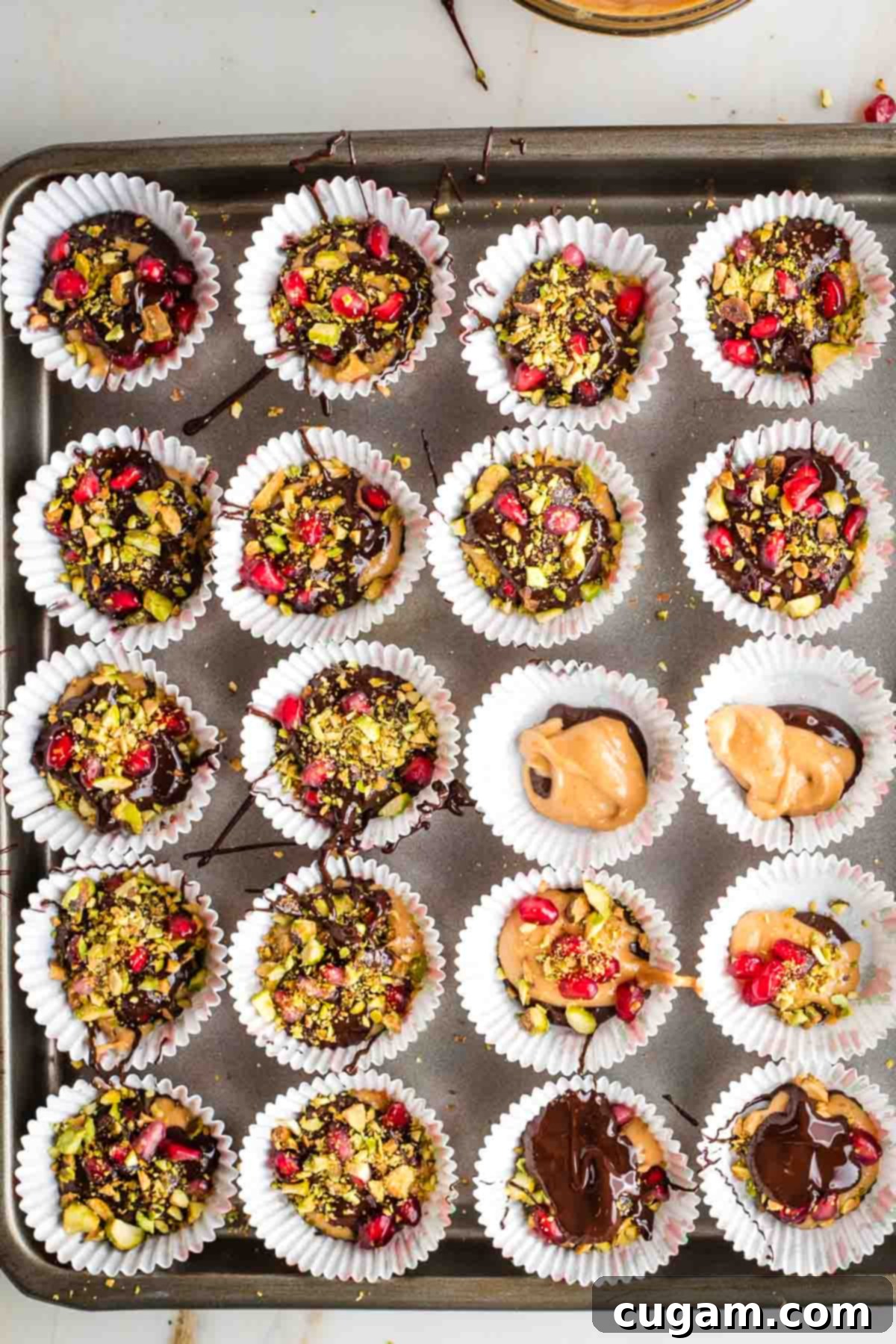 Rimmed baking tray with mini cupcake liners filled with chocolate that has been stuffed with peanut butter and sprinkled with nuts and pomegranate seeds.