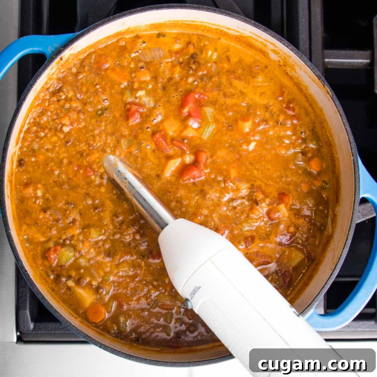 Immersion blender in the center of a dutch oven filled with chunky vegan lentil soup, actively pureeing a portion for creaminess.