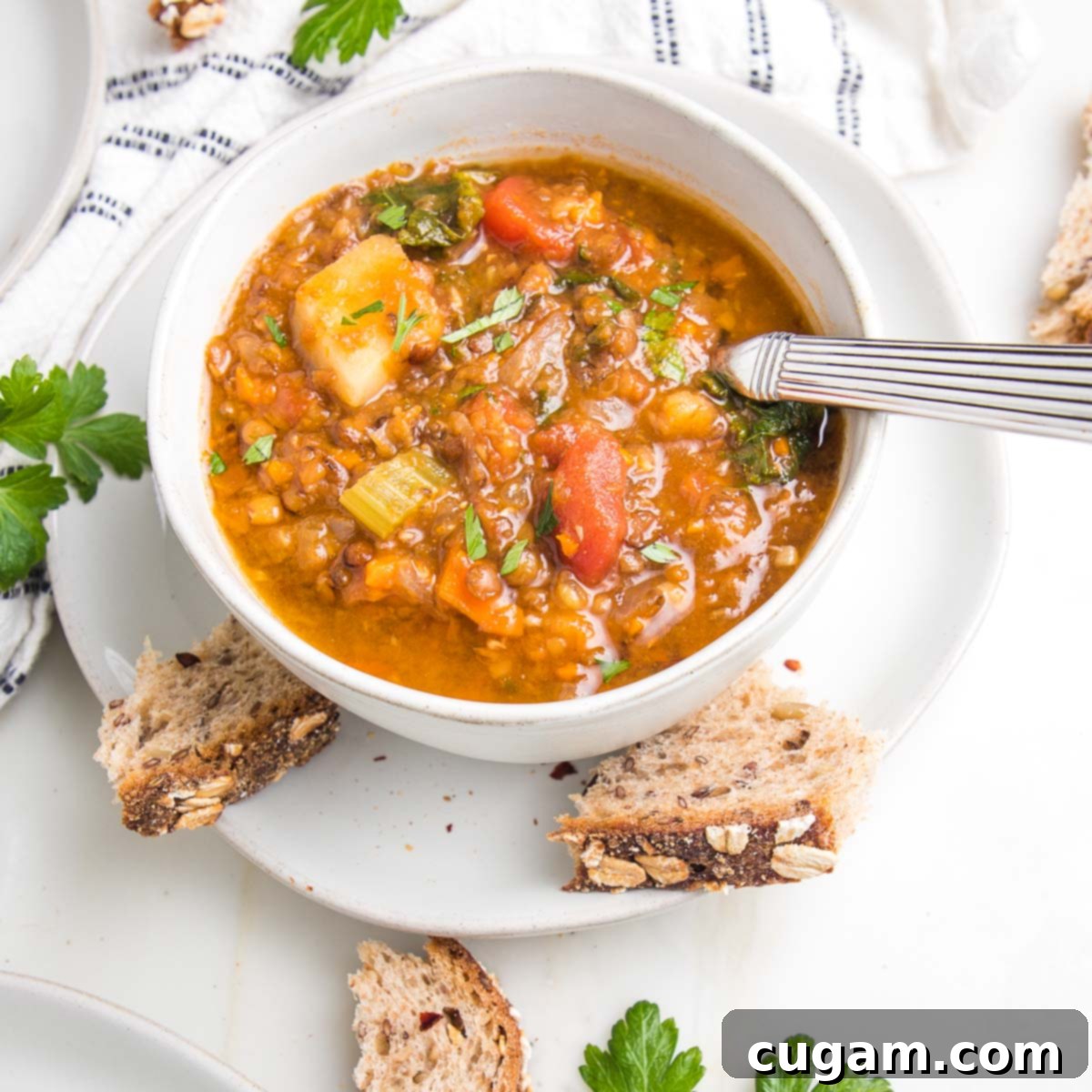 Chunky lentil and vegetable soup with crusty chunks of bread, fresh sprigs of parsley and a white with navy striped dish towel. A hearty and inviting scene.