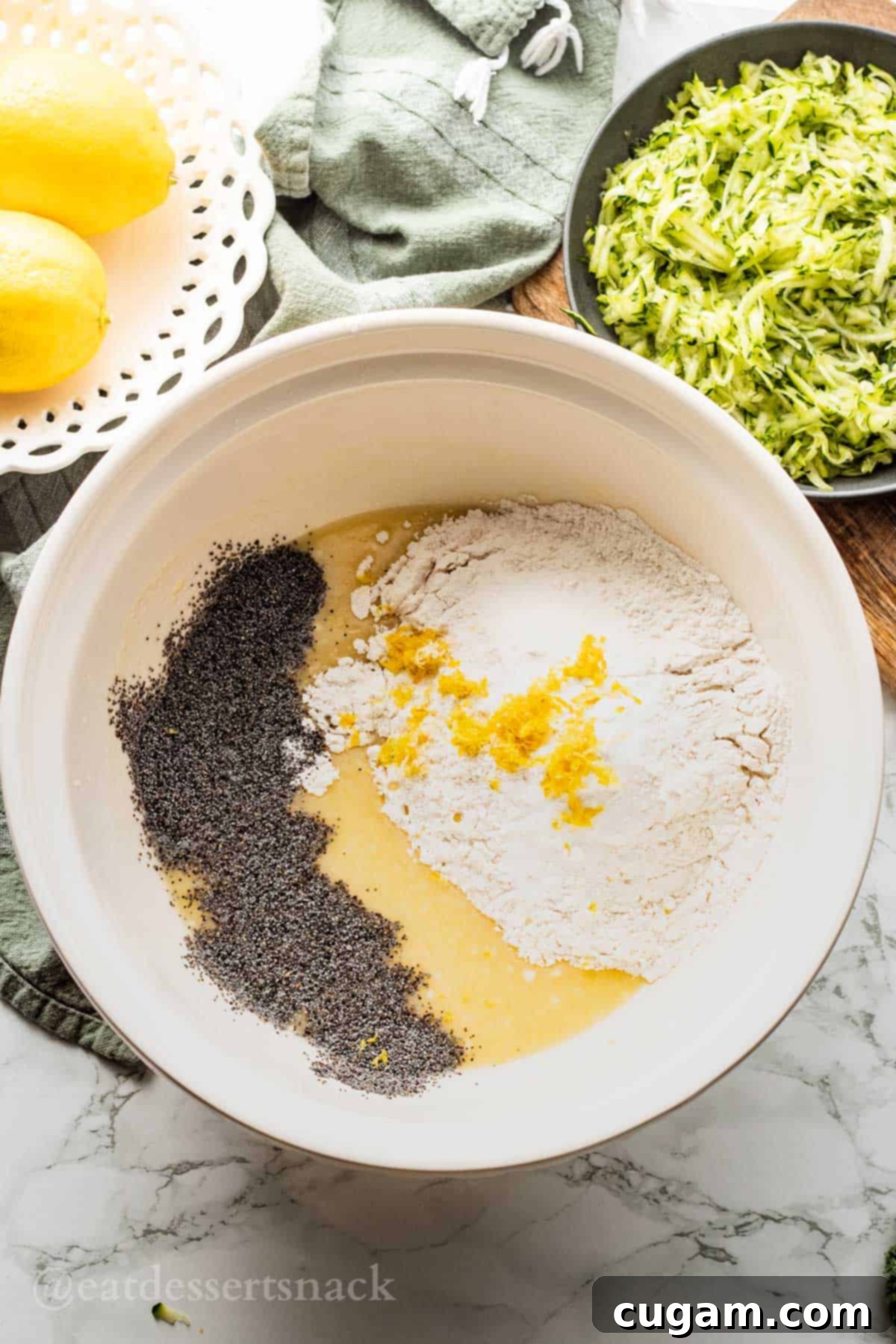 Flour, lemon zest, and poppyseeds on top of mixed wet ingredients in white porcelain mixing bowl.