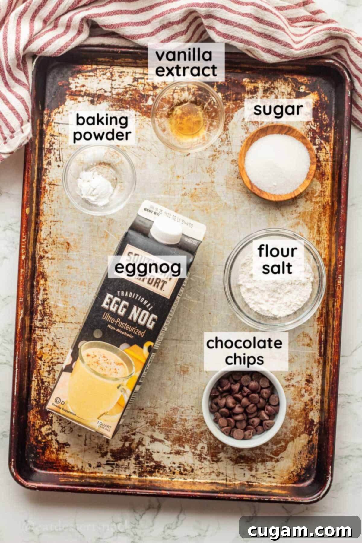 Labeled ingredients for eggnog mug cake in glass bowls on metal baking sheet, ready for preparation.