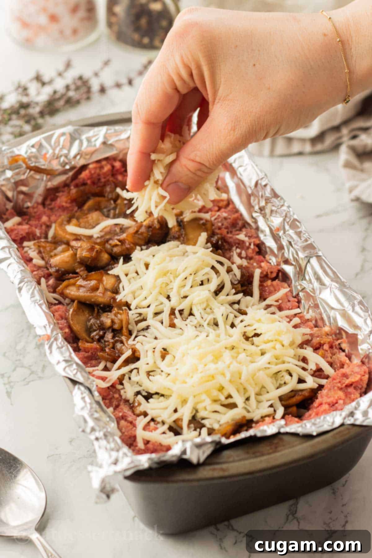 Hand sprinkling cheese onto cooked onions and mushrooms in raw beef in metal pan.