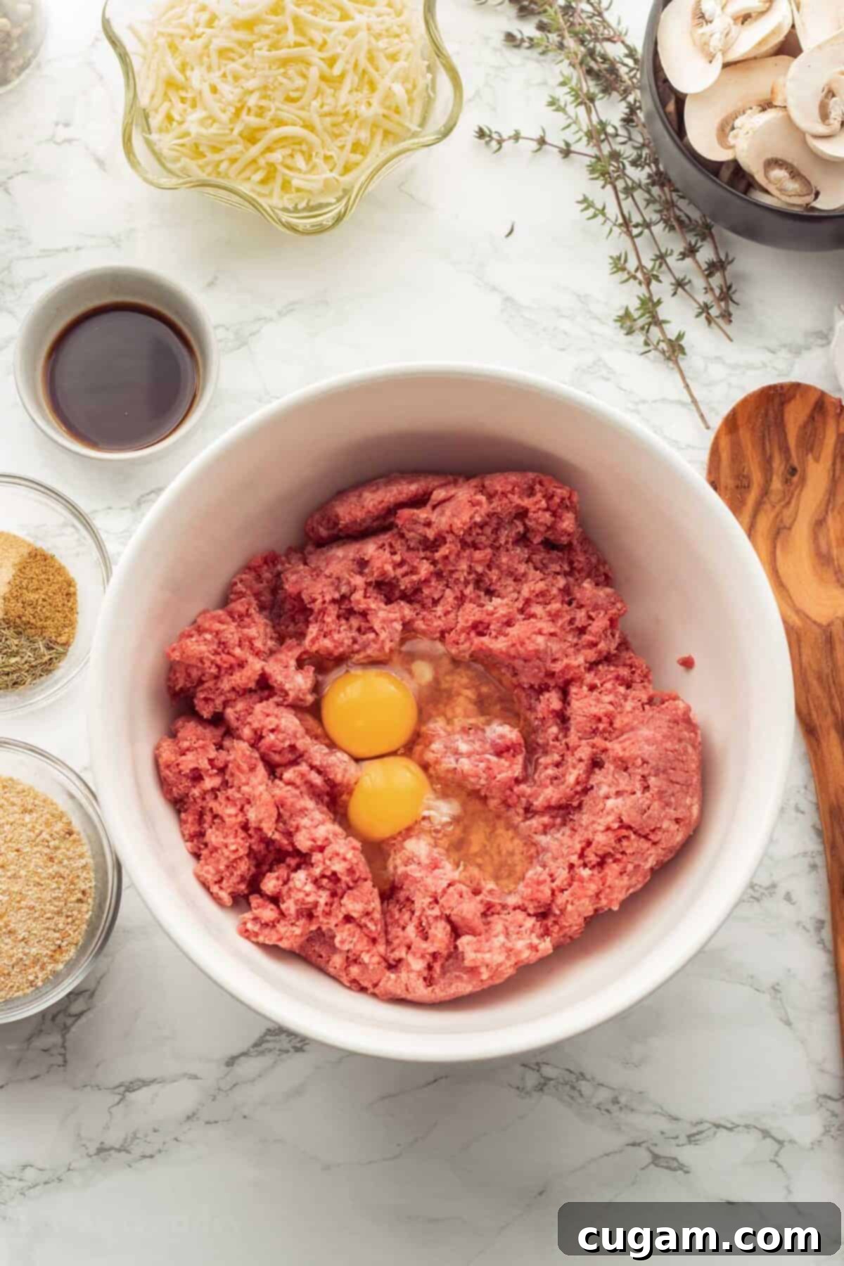 Raw ground beef in white bowl with 2 eggs on marble countertop with glass bowls of spices and wood spoon.
