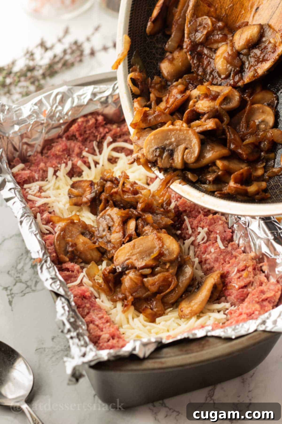 Pouring cooked mushrooms and onions into raw beef, over a layer of cheese.