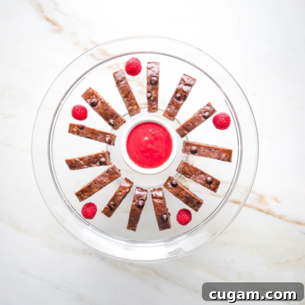 Thinly sliced chocolate zucchini brownies arranged in a circle around a small bowl of vibrant raspberry sauce, perfect for dipping.
