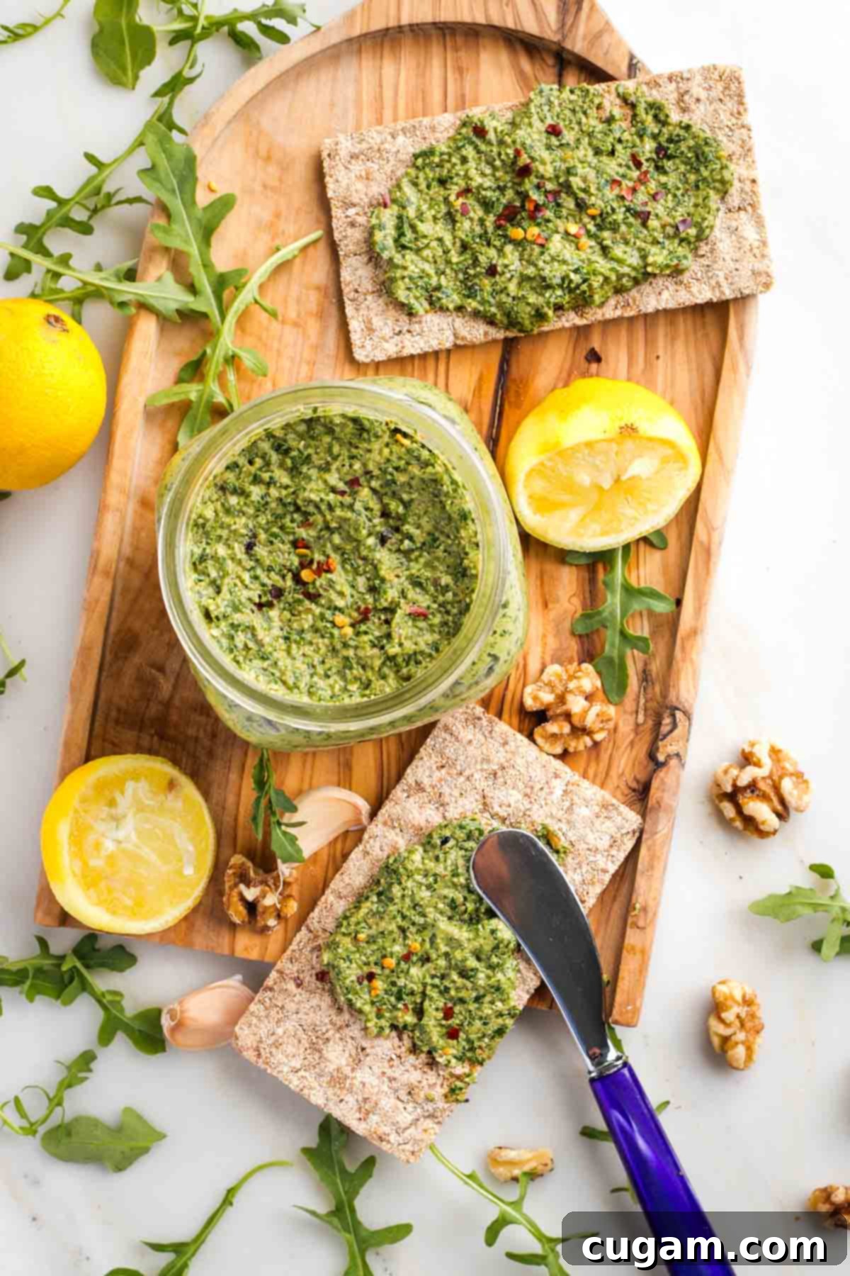 Vegan Arugula Walnut Pesto 7 Jar of pesto surrounded by lemon, walnuts, arugula and garlic with long crackers spread with pesto.
