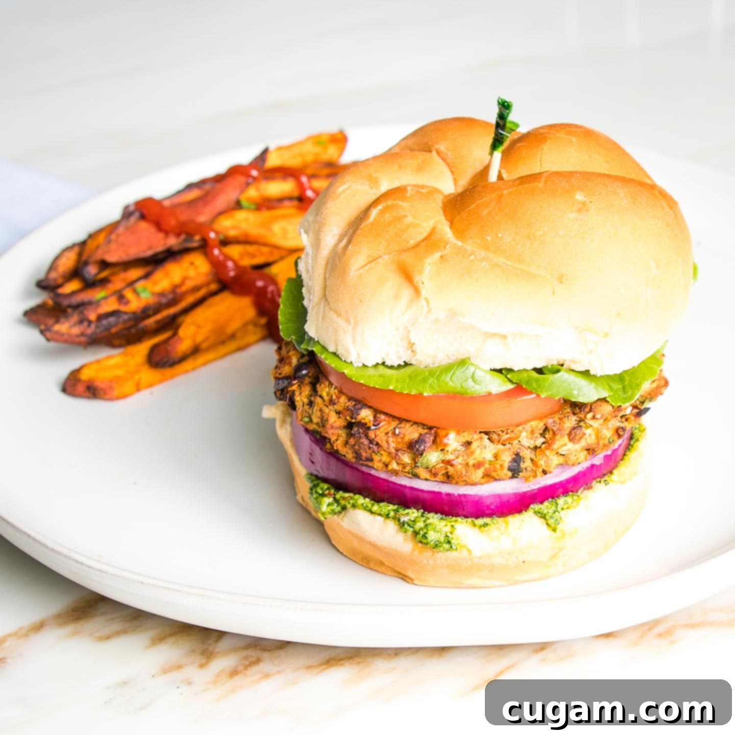 Veggie burger on a bun with green arugula aioli, purple onion, lettuce and tomato on a bun with sweet potato fries on the side of a round white rimmed plate.