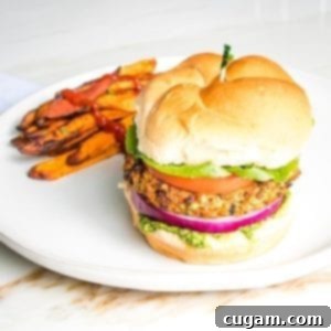 Veggie burger on a bun with green arugula aioli, purple onion, lettuce and tomato on a bun with sweet potato fries on the side of a round white rimmed plate.