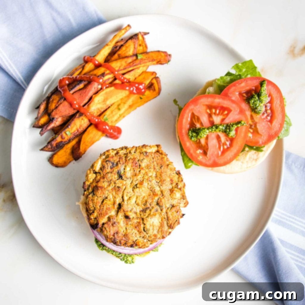 White bean burger on a bun with lettuce and tomatoes and purple onion and arugula aioli plus sweet potato fries drizzled with ketchup on a round white plate. 