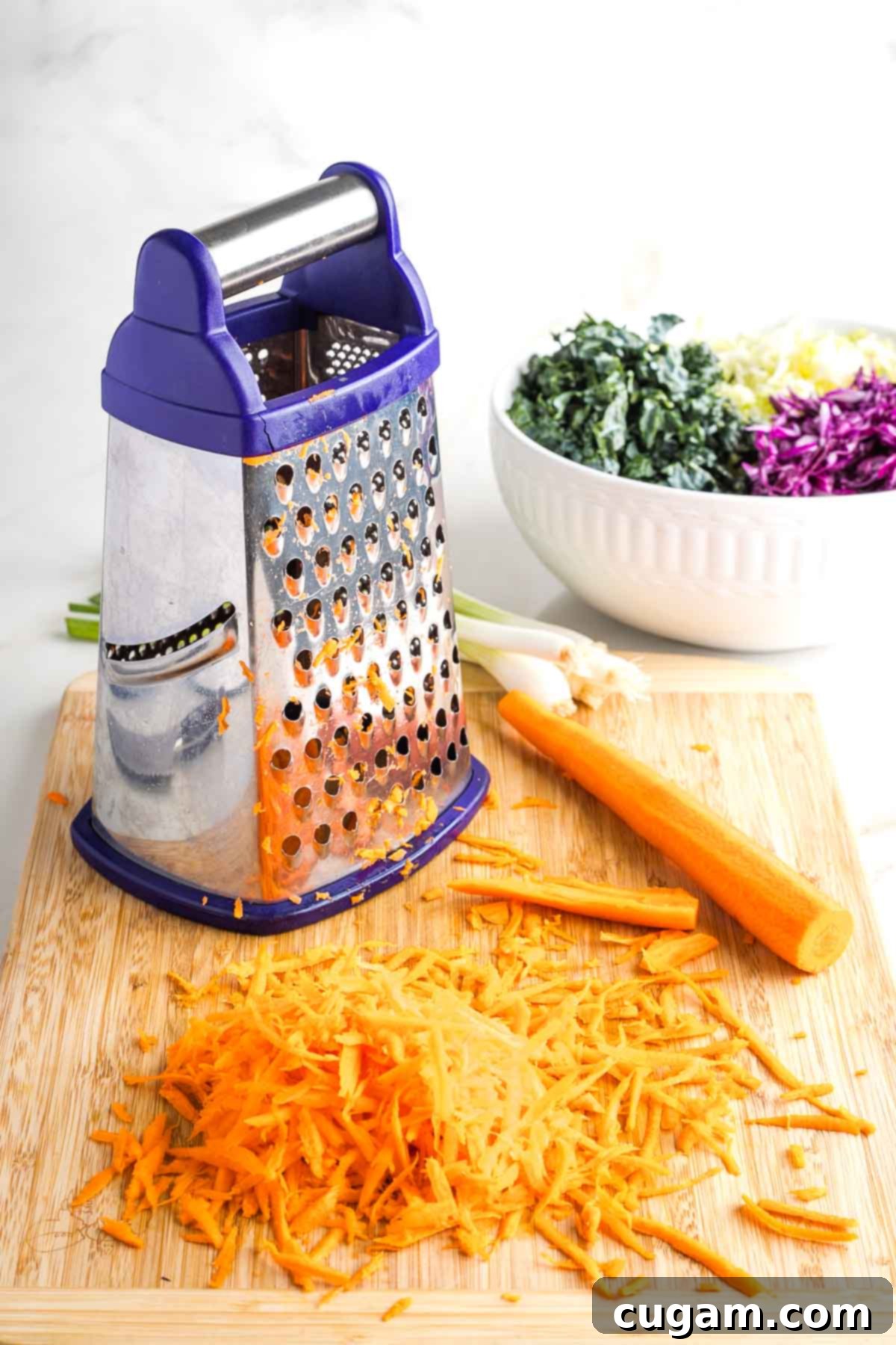 Box grater, shredding carrots with a bowl of shredded cabbage in the background. 