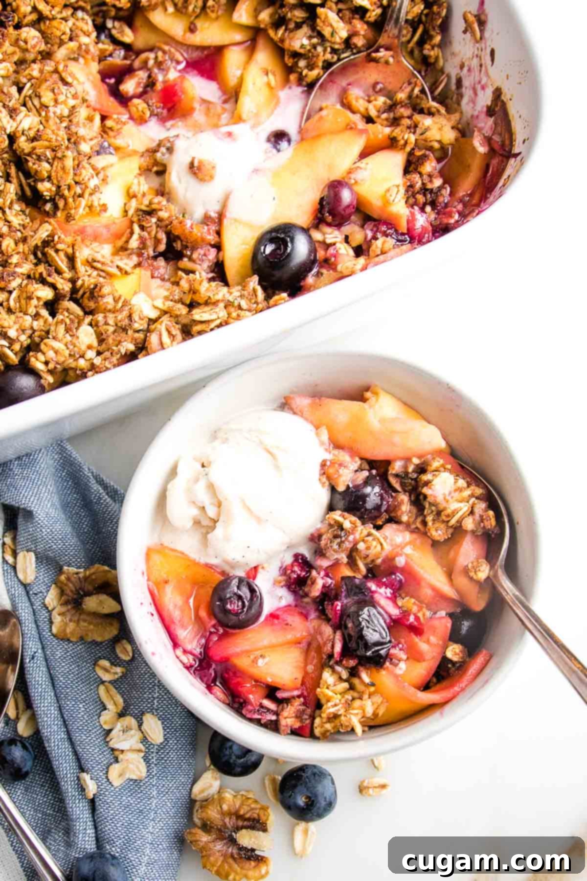 Rectangle white casserole dish filled with fruit and berry crisp and some vanilla ice cream. In front is a small white bowl with peach and blueberry crisp and a scoop of vanilla ice cream. There is a blue dish towel and some walnuts, oats and blueberries on the white countertop as well.