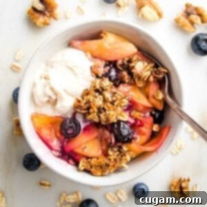 White bowl filled with peach and blueberry crisp--oatmeal and nut topping and a scoop of vanilla ice cream. The white countertop is strewn with almonds, fresh blueberries and oats.