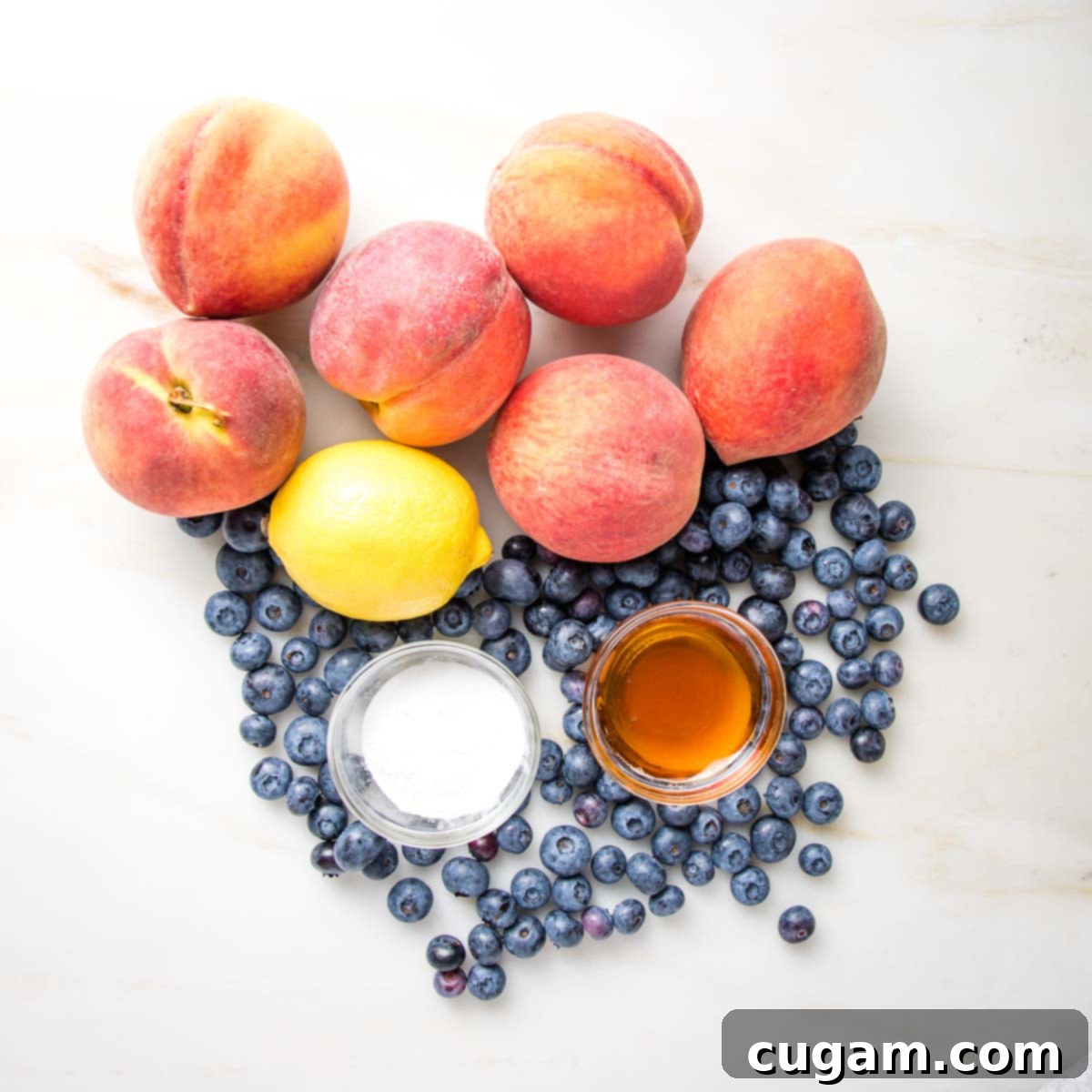 Peaches, blueberries, and a lemon on a white countertop. Some arrowroot powder and maple syrup in a little glass bowls.