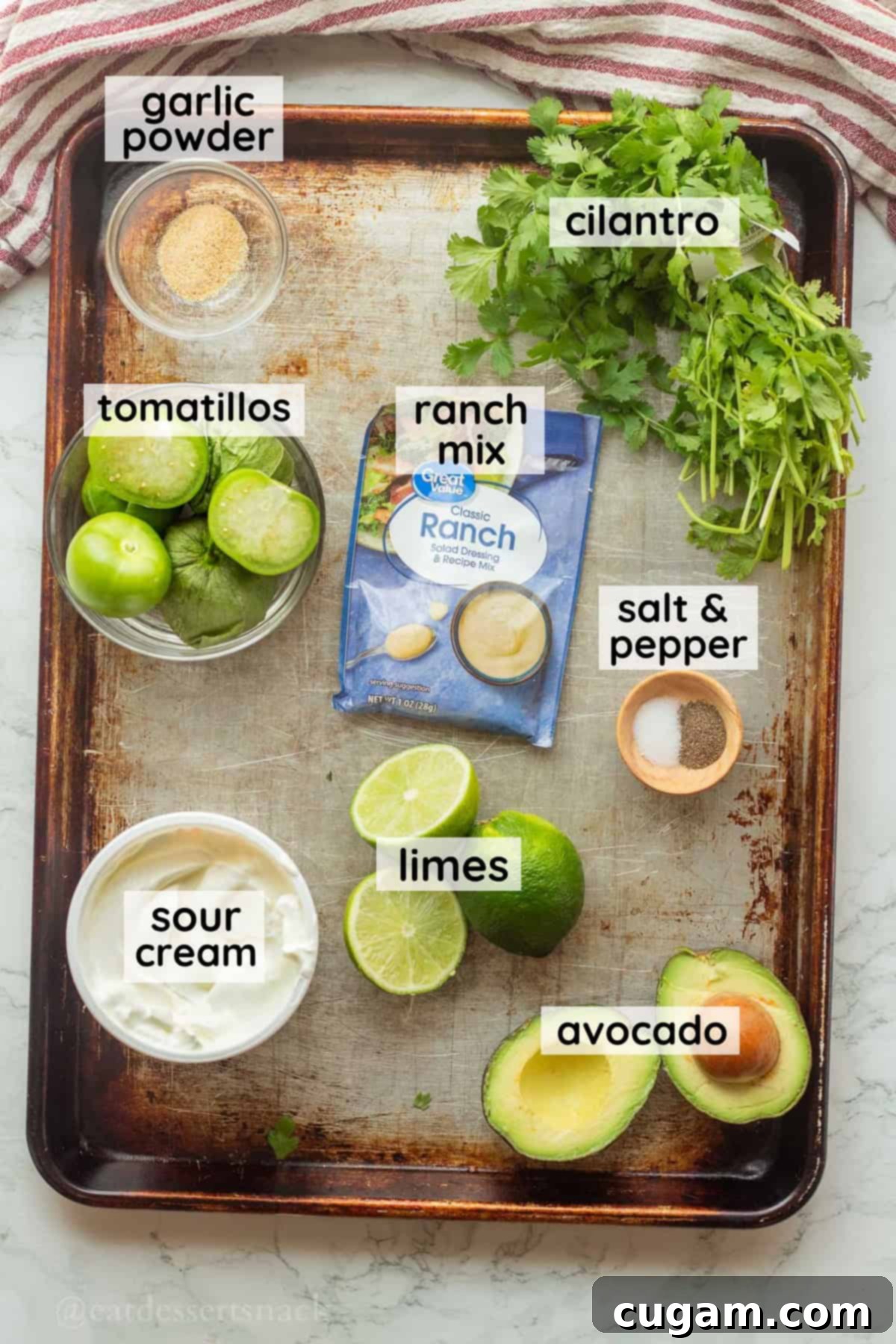 Individual glass bowls containing perfectly ripe avocados, quartered tomatillos, halved limes, and a packet of ranch dressing mix, all neatly arranged on a metal baking sheet for a visual guide to the key ingredients.