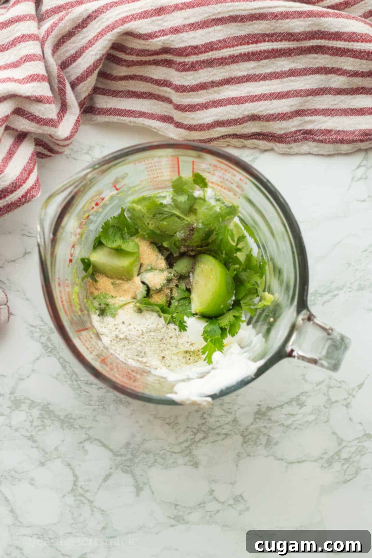 A glass measuring cup filled with vibrant green ingredients for avocado lime ranch, including chopped tomatillos, lime halves, creamy avocado, sour cream, and various seasonings, all ready to be blended.