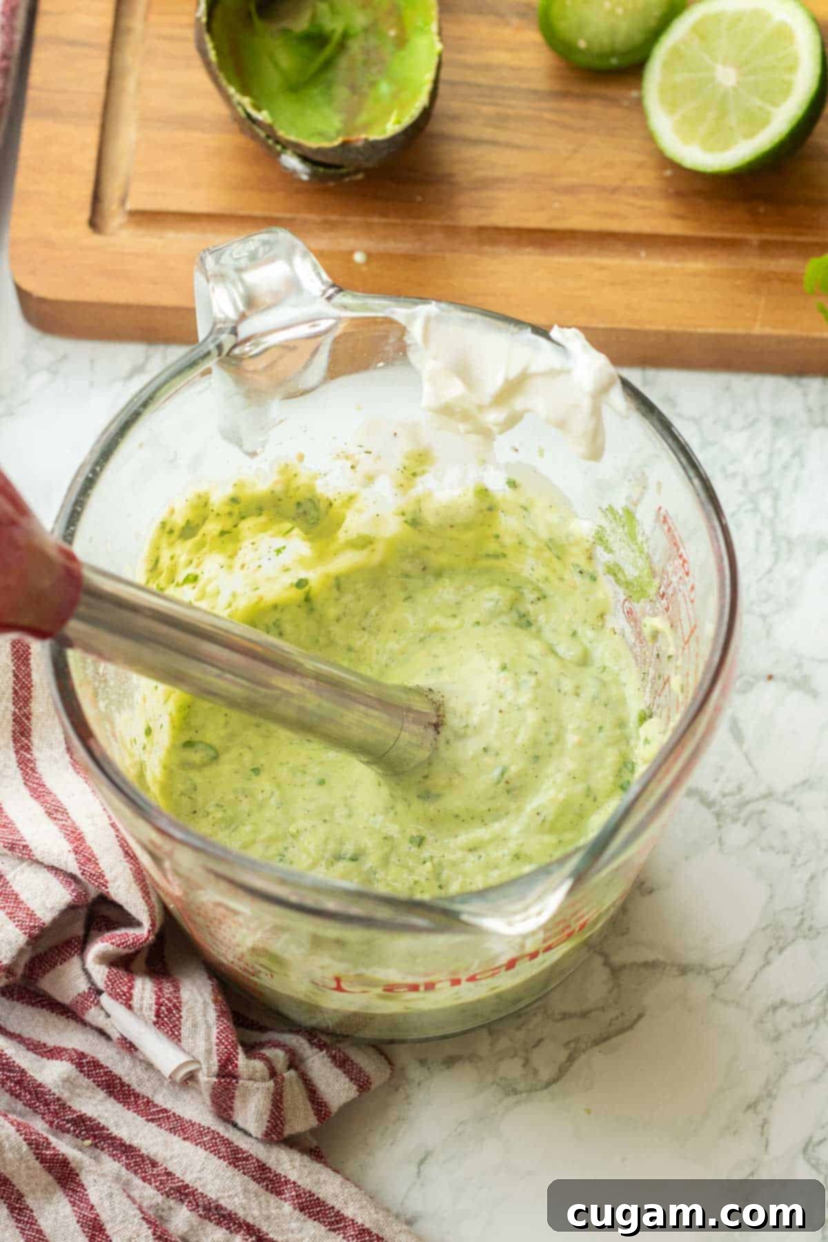 An immersion blender actively mixing the creamy green salad dressing ingredients in a tall glass measuring cup, creating a smooth and uniform consistency.