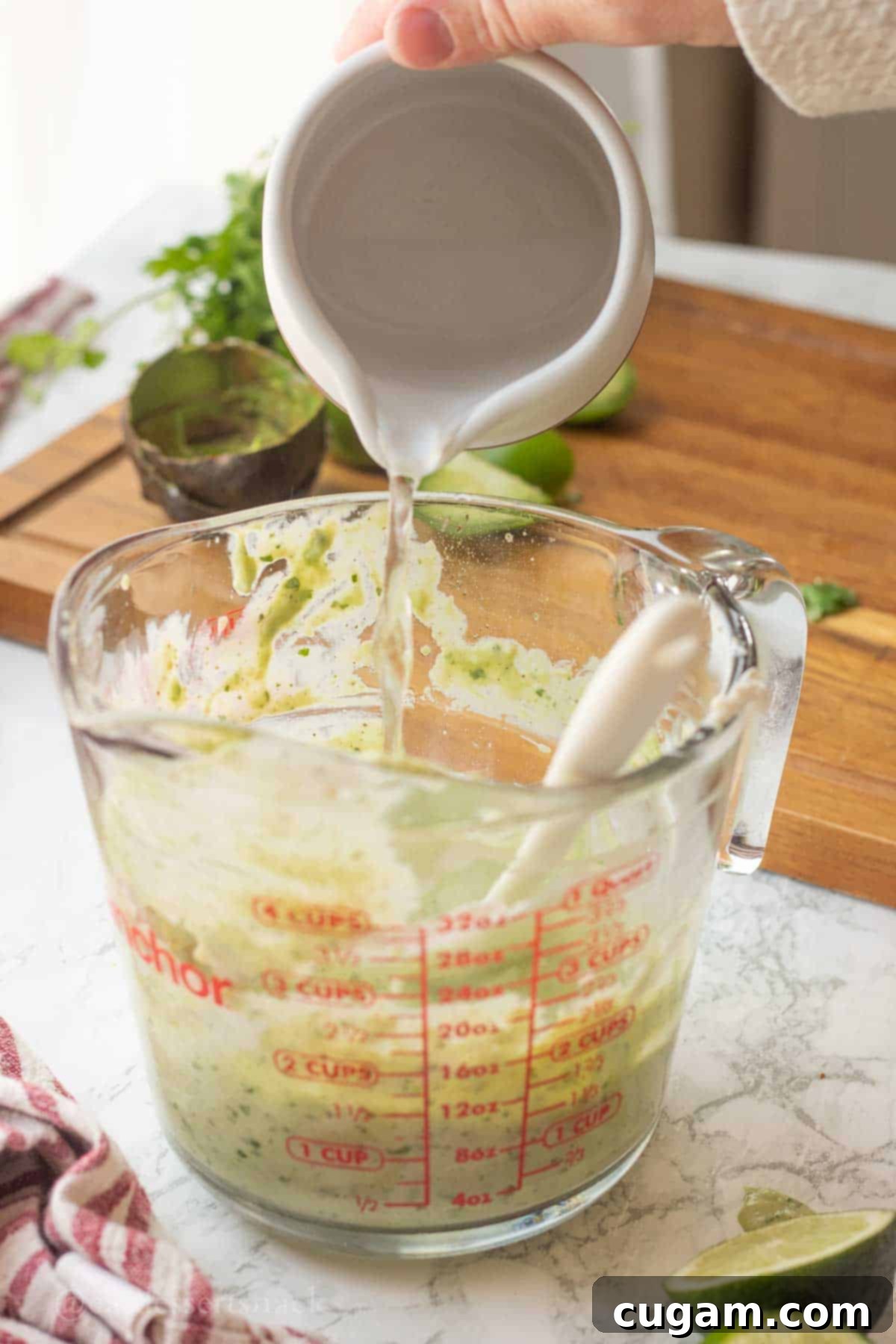 A hand pouring water into a measuring cup filled with the blended green salad dressing ingredients, demonstrating the step to adjust consistency.