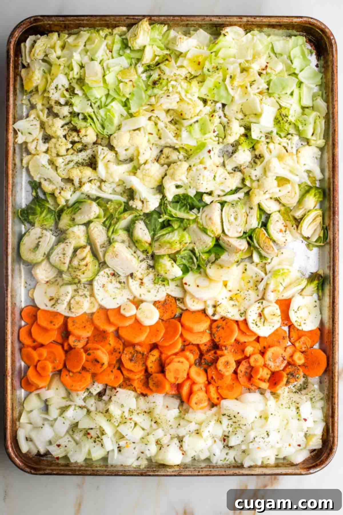 Rows of chopped root vegetables and cruciferous vegetables on a large baking tray, drizzled with oil and sprinkled with Italian spices and red pepper flakes.