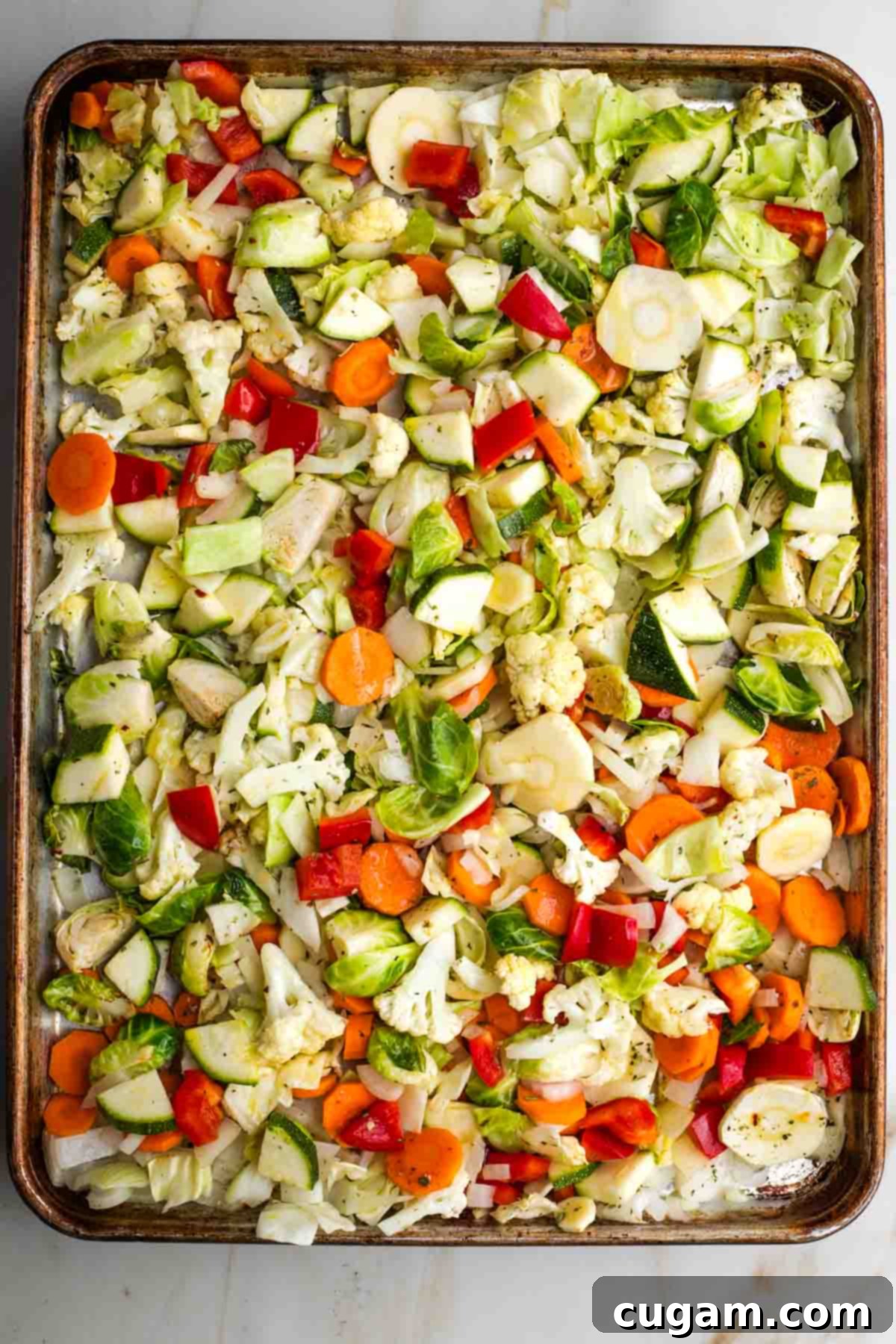 Colorful assortment of chopped vegetables including zucchini and red bell pepper added to a large rimmed baking sheet with pre-roasted veggies.
