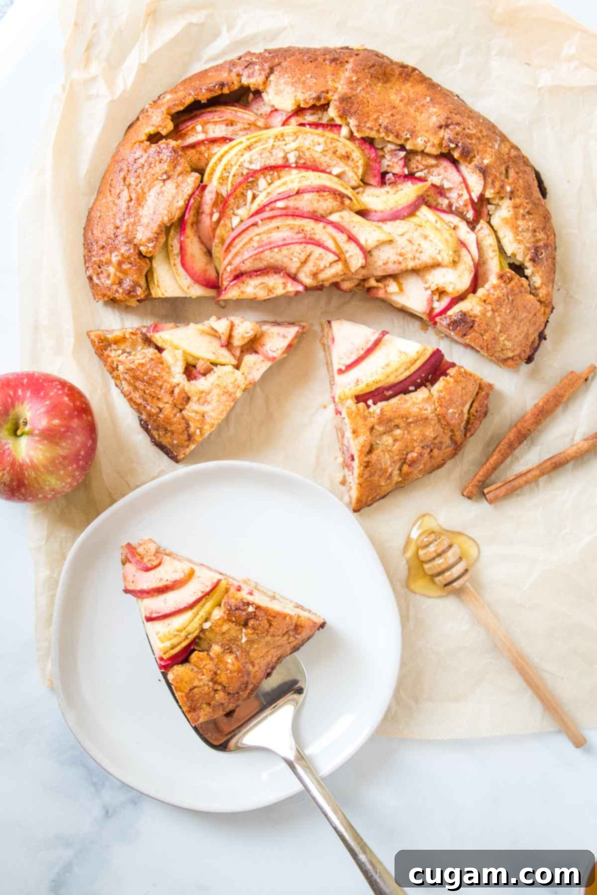 Free form apple galette, half of it sliced. One piece is on a plate. There is an apple, some cinnamon sticks and a dollop of honey on the parchment paper that the galette is also on.