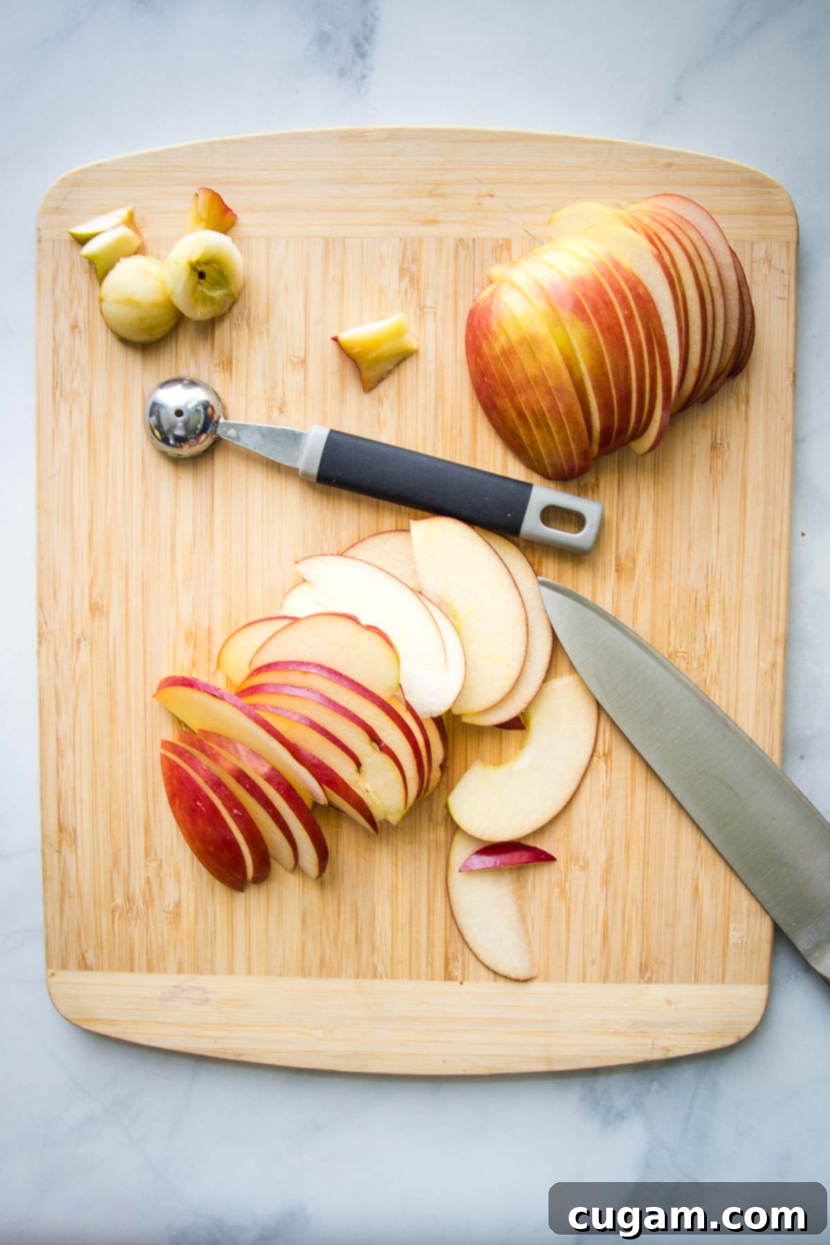 Wooden cutting board with red apple sliced thinly in two halves. A melon baller is in the center and the core in the upper left corner.