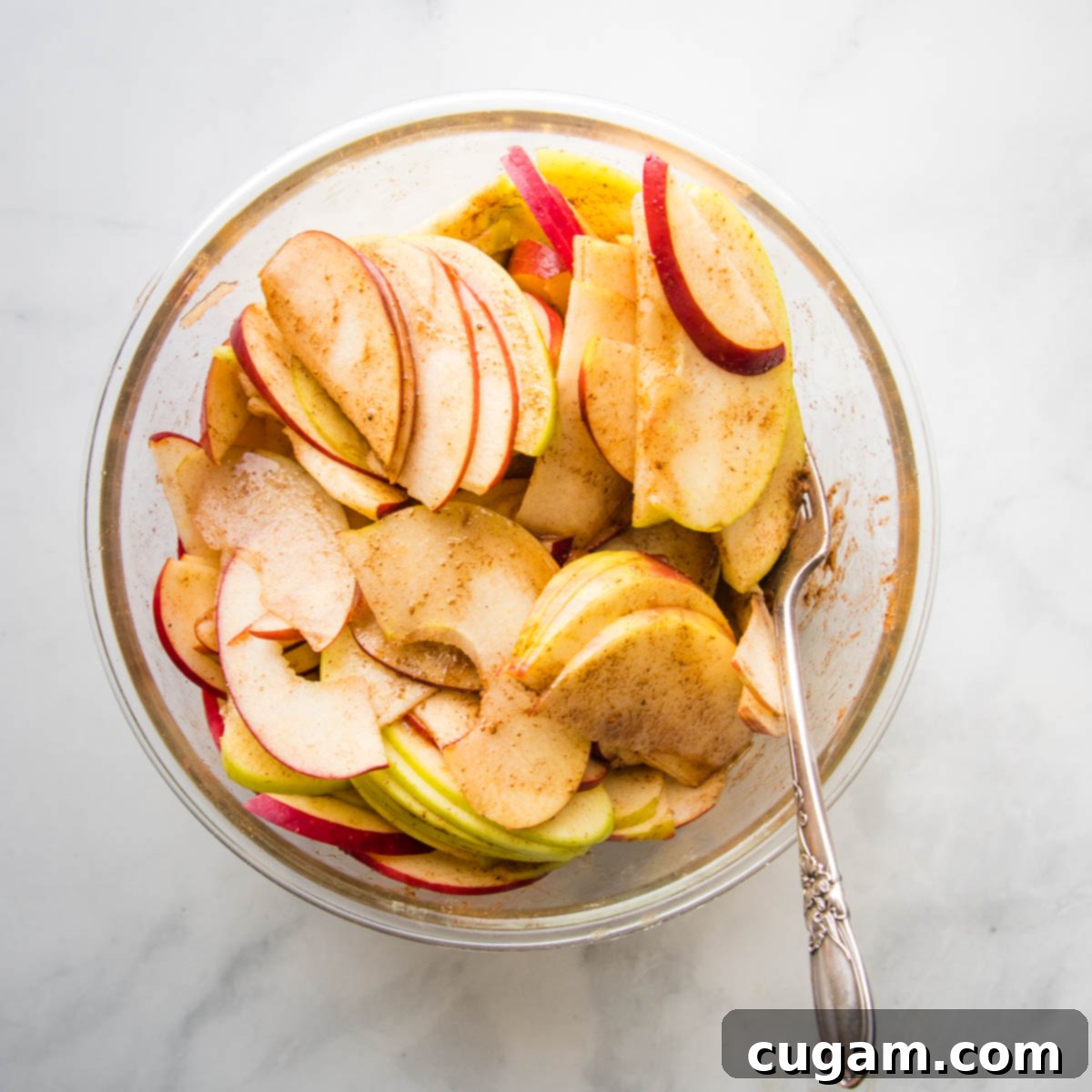 Green and Red Sliced Apples, mixed with apple pie spices, in a large glass bowl.
