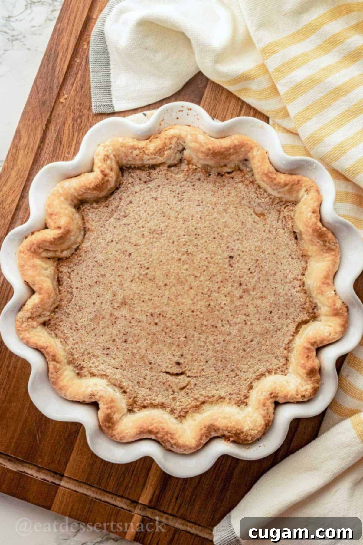 Baked banana pie in white fluted pie pan on wood cutting board with yellow striped dish towel.