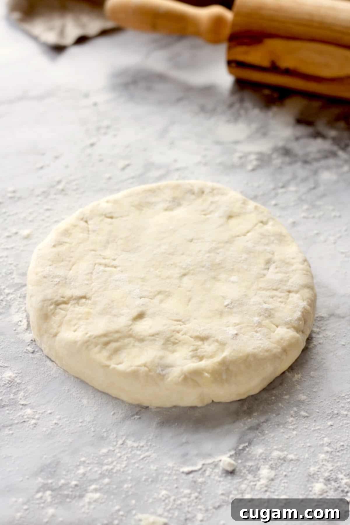 Disc of raw cream cheese pie dough on marble countertop with wooden rolling pin.