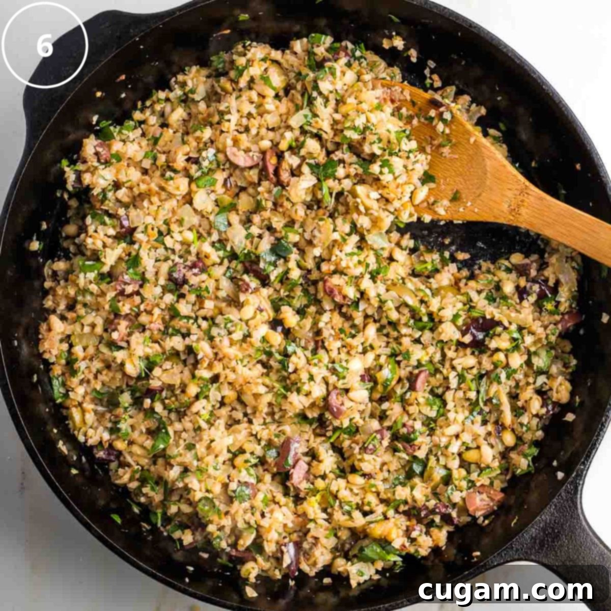 Vibrant Mediterranean Cauliflower Rice 9 Step 6: Wooden spoon stirring mediterranean cauliflower rice in a cast iron skillet with olives, fresh herbs and pine nuts.