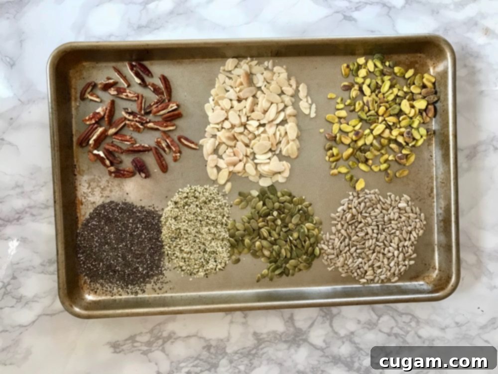 An array of raw nuts and seeds laid out on a rimmed baking sheet, including pistachios, almonds, pecans, pepitas (pumpkin seeds), sunflower seeds, chia seeds, and hemp seeds, ready for toasting.